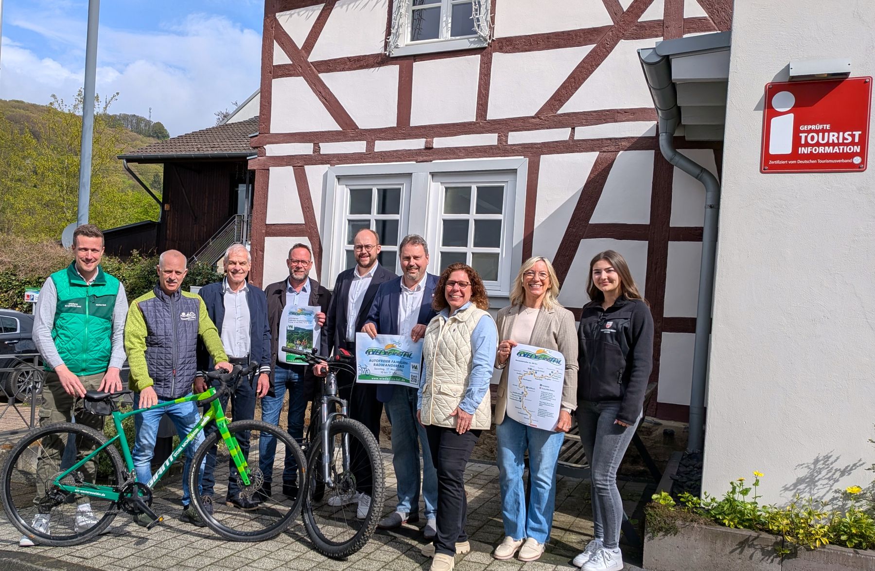 Eine Gruppe von neun Personen steht lächelnd vor einem Fachwerkgebäude mit einem roten Schild der Touristeninformation. Zwei Personen halten Schilder, und ein grünes Mountainbike ist davor geparkt.
